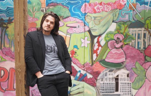 Riley Brenes stands in front of a mural that will be at this weekend’s Festival of Flowers in Cathedral Square. The mural was previously on display in the courtyard at Strickland Youth Center. It features a portrait of Sam Lackland, Mobile scenes and bright pink azaleas. 